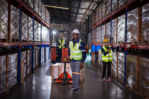 Image depicts a man in a yellow high-visibility vest pulling a cart of palettes through a well lit warehouse.