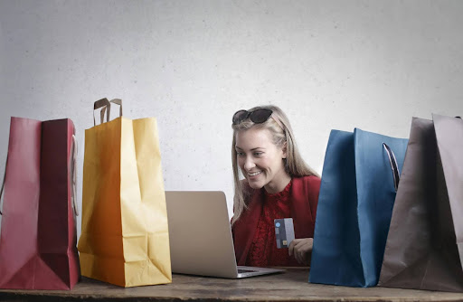 A blonde woman in a red sweater with sunglasses on her head is holding a credit card and shopping on a silver laptop. On the wooden table she is sitting by, colorful shopping bags surround her in red, yellow, blue, and gray. Behind her is a gray speckled wall.